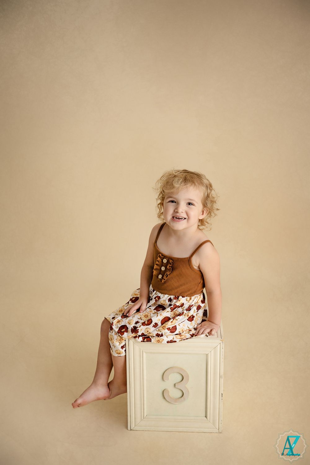 Smiling toddler in brown tank and floral skirt sits on decorative white cube.
