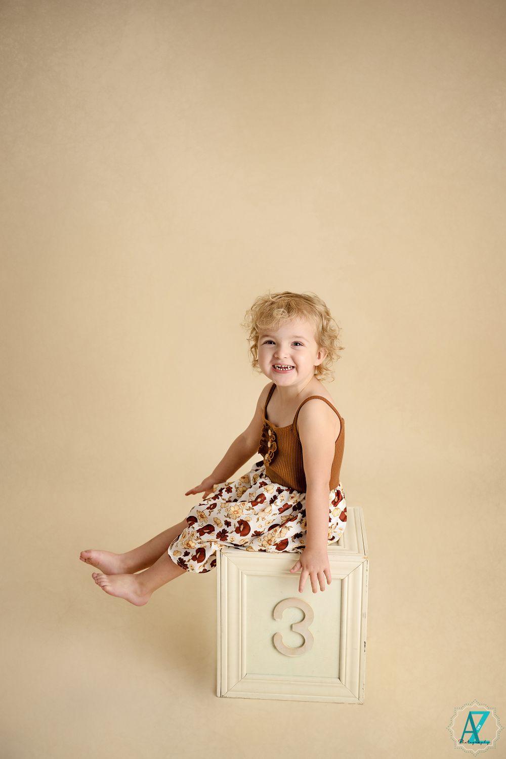 Laughing toddler sits on white numbered block wearing brown and floral outfit.