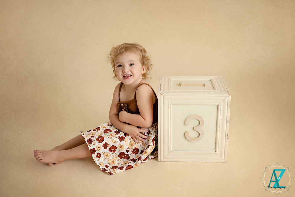 Cheerful toddler in floral dress sits beside white numbered cube against beige background.