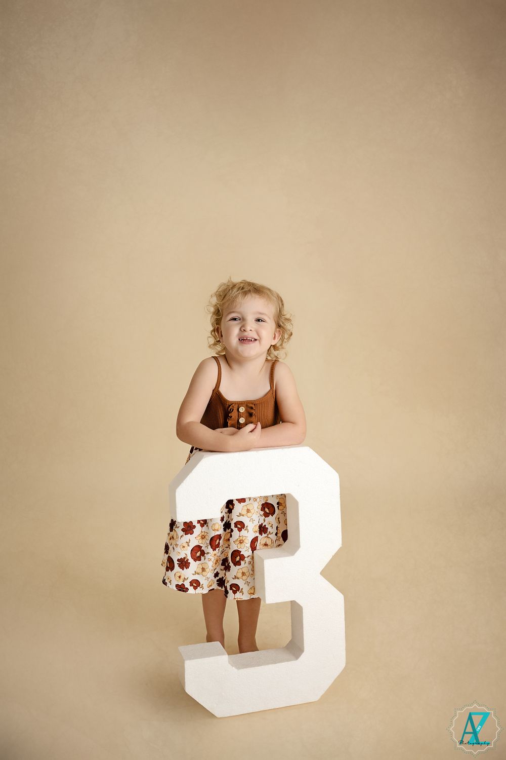 A small child wearing a brown top and floral skirt stands on a large white number 3 against a beige backdrop.