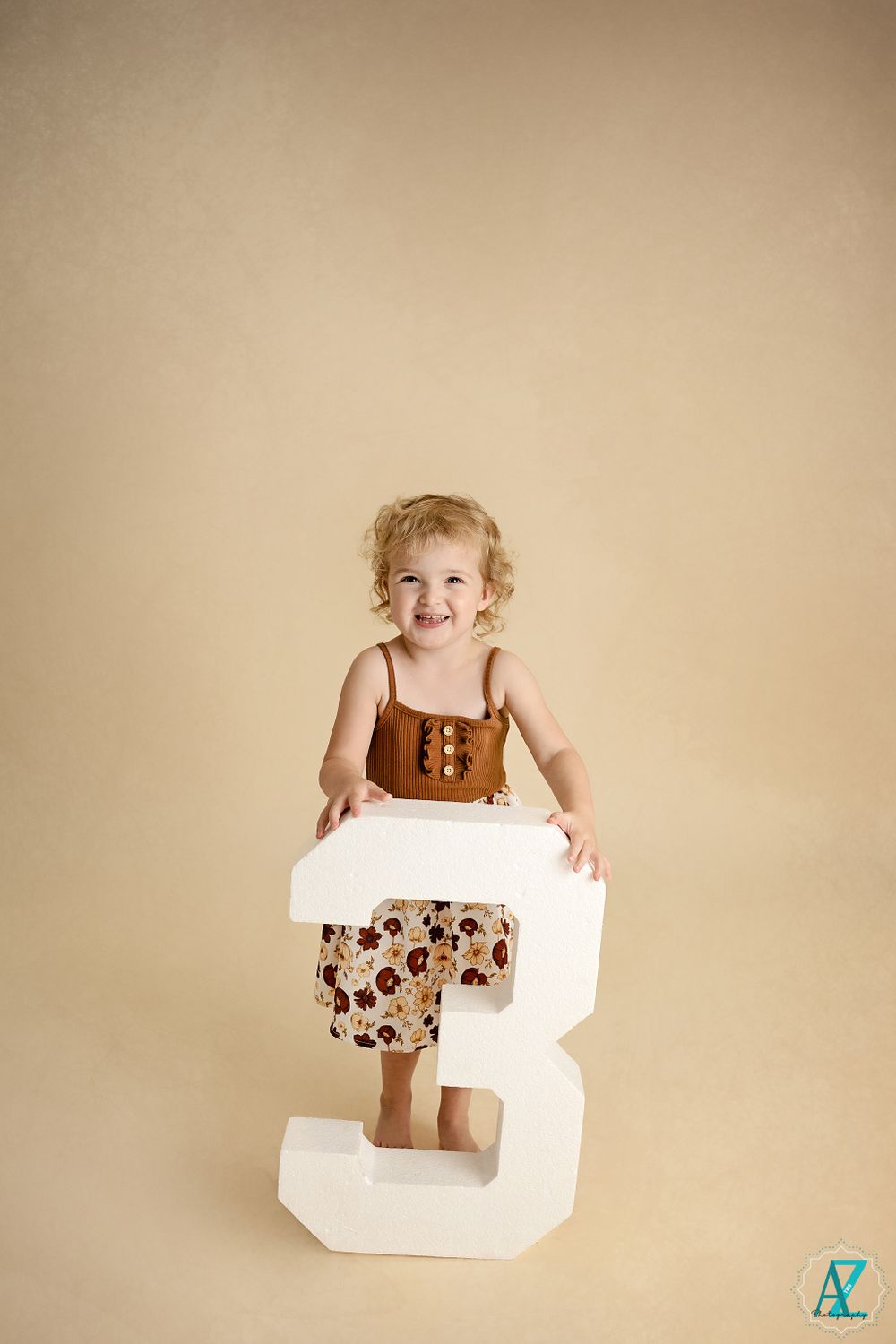 A barefoot toddler in brown and floral outfit poses playfully on a white number 3 prop.