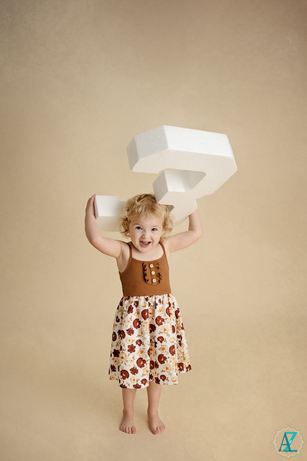 A joyful child holds up a large white number 3 while wearing a brown sleeveless top and patterned skirt.