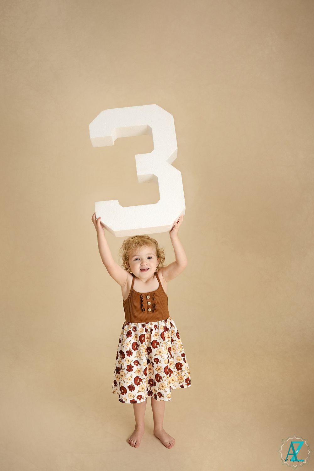 A young child raises a large white number 3 overhead while standing barefoot against beige background.
