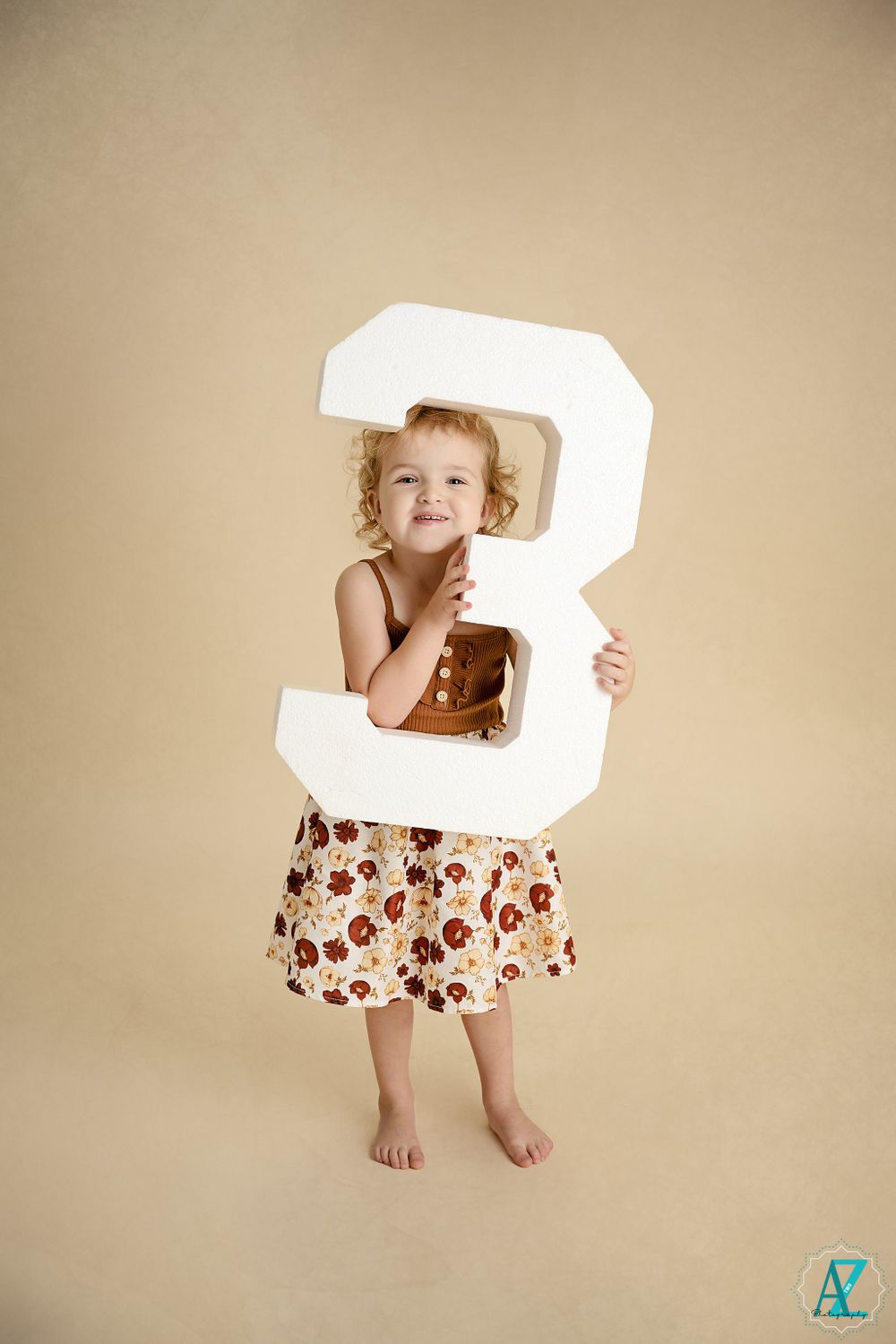 A child peeks through a large white number 3 while wearing a brown top and floral pattern skirt.