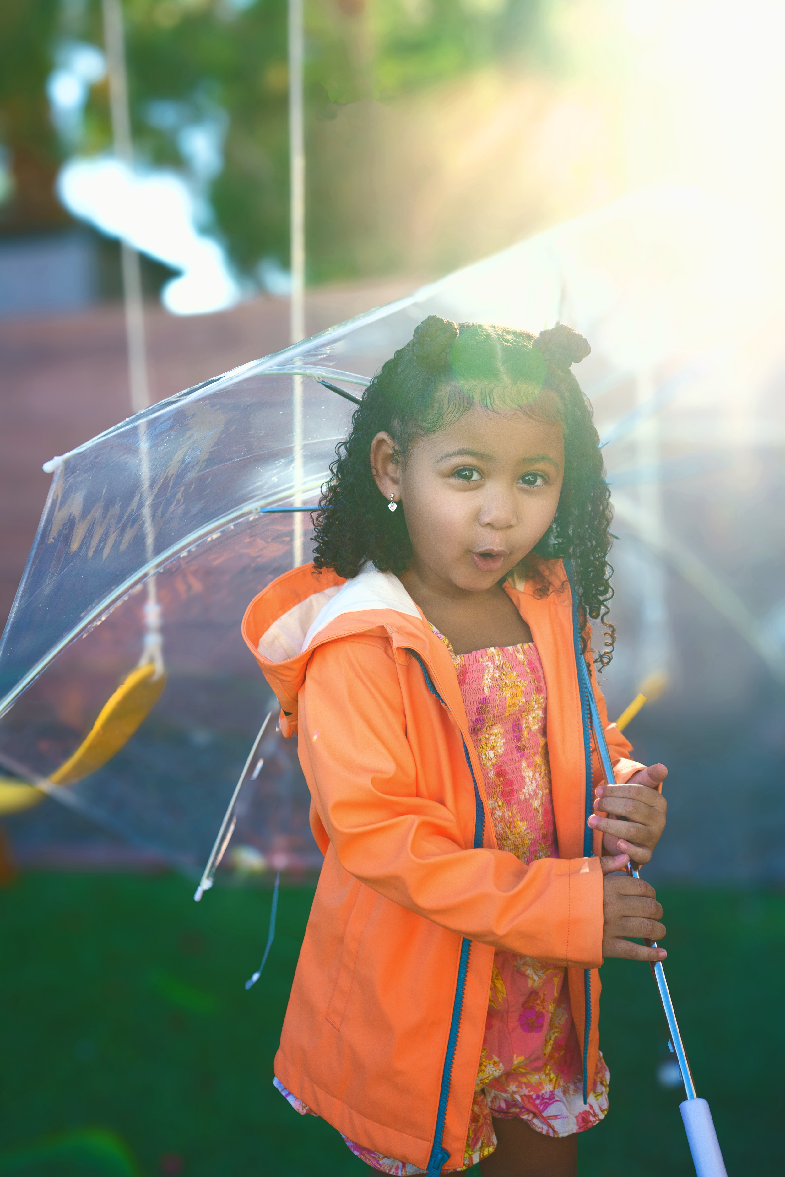 Child in orange jacket holding clear umbrella outdoors on sunny day.