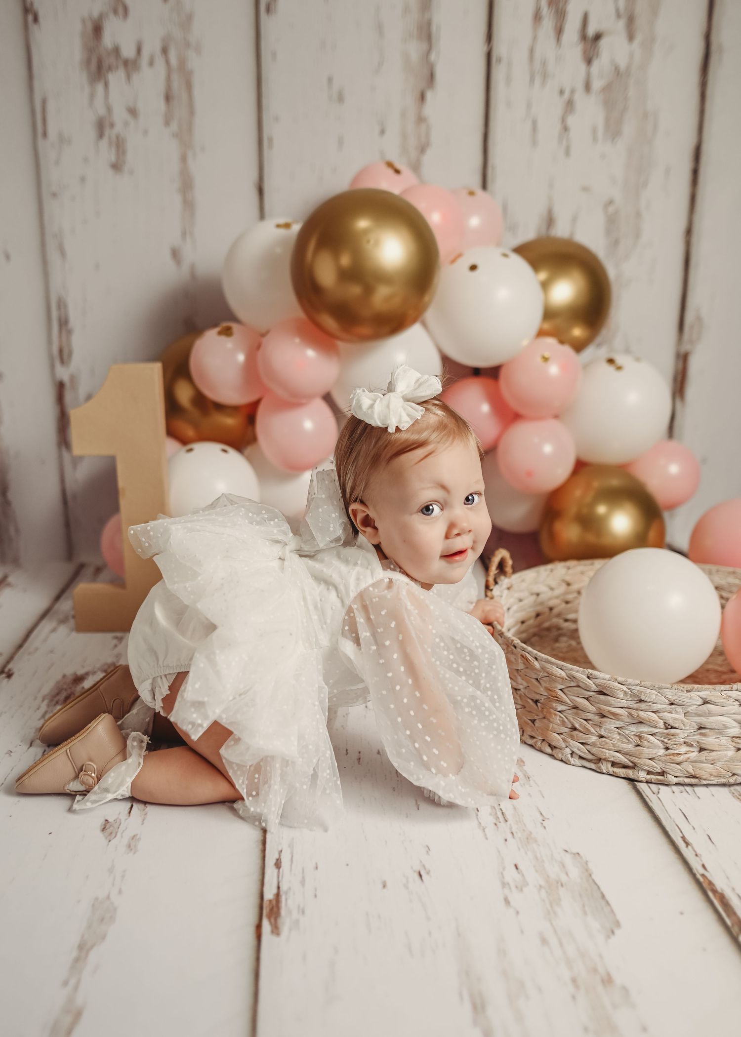 Baby in white lace dress with gold and pink balloon decorations in vintage setting.