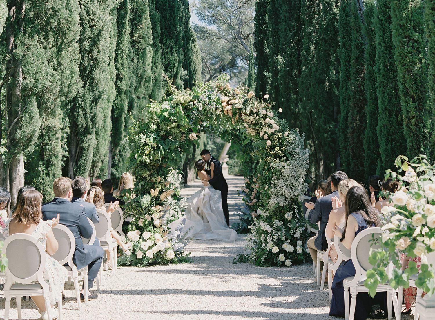 Beautiful outdoor wedding ceremony set up with floral arch and white chairs along cypress-lined path.
