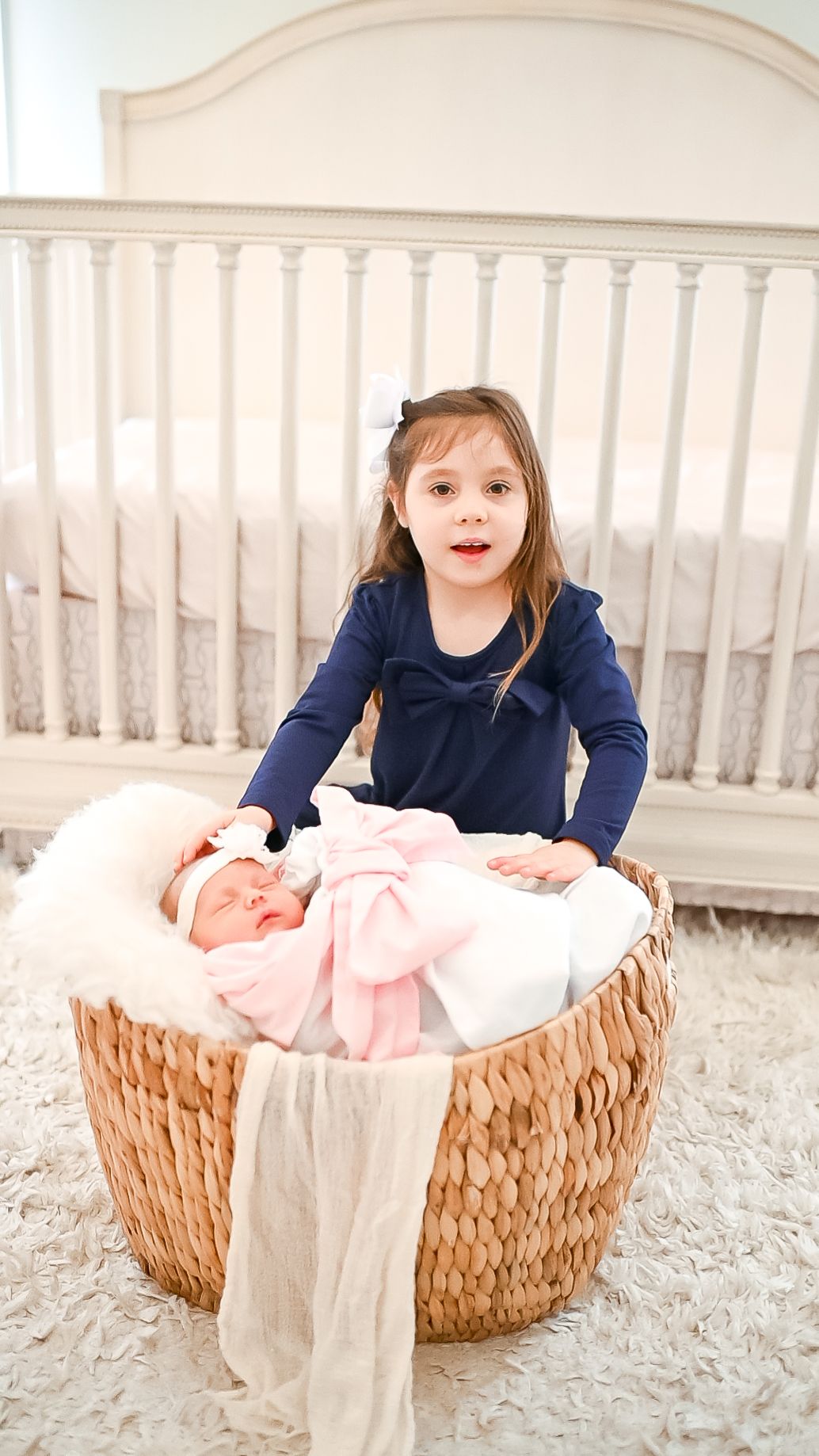 sister wearing a blue dress with a bow touching her newborn baby sister on the head taking portraits