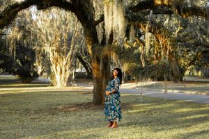 Figure in patterned dress standing under sunlit oak trees with hanging moss.