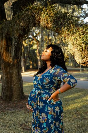 Maternity photoshoot in a park featuring a blue floral dress against Spanish moss-draped trees in golden sunlight.