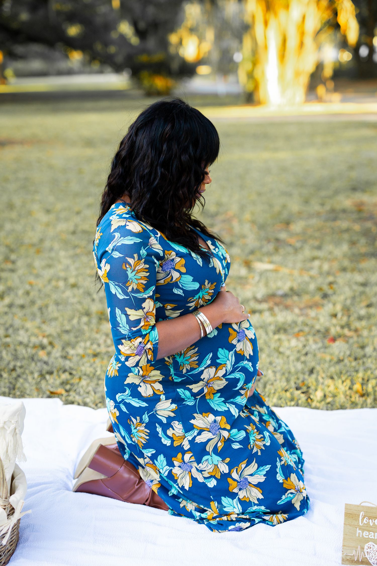 Side view of pregnant woman in floral dress sitting on white blanket during outdoor photoshoot.