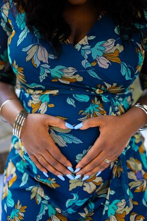 Close-up of hands forming heart shape on baby bump in blue floral maternity dress.