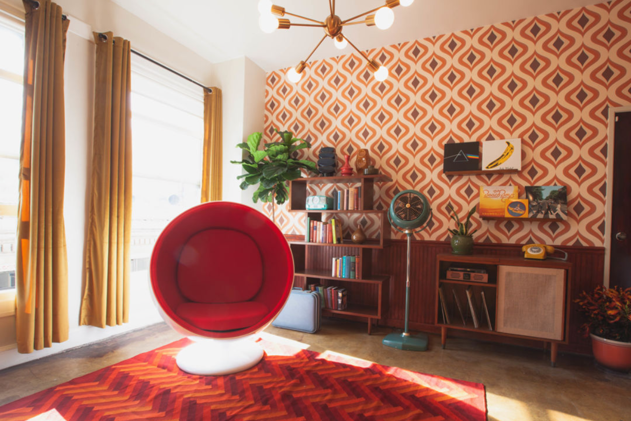 Mid-century modern room with orange geometric wallpaper, red egg chair, and wooden shelving unit.