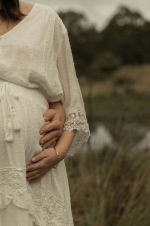 Close-up of hands cradling a pregnant belly in a white lace dress against a natural backdrop.