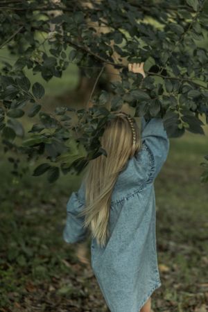 Long-haired child in denim dress reaching up to touch tree branches in a garden setting.