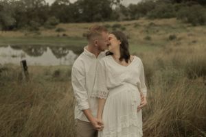 A romantic moment between a couple in white clothing standing in a grassy field near water.