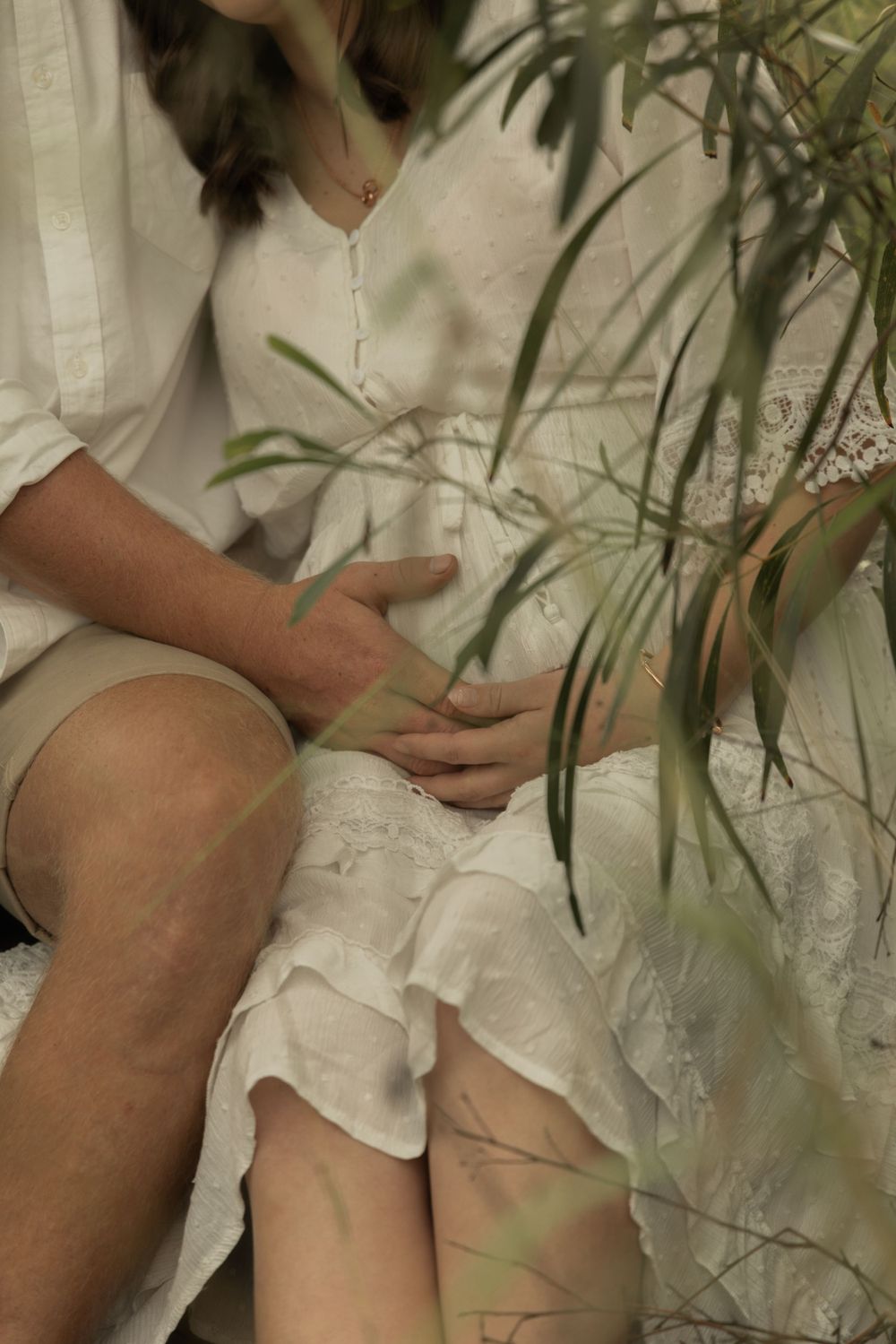Artistic detail shot of someone sitting in a white dress with green foliage in foreground.