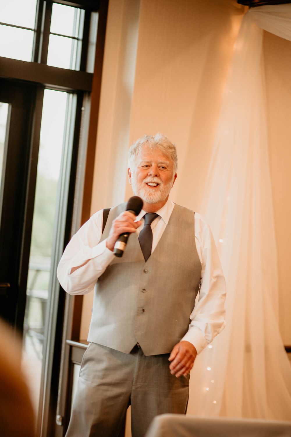 Older man with white beard giving a wedding speech while holding a microphone, wearing a gray vest and tie.
