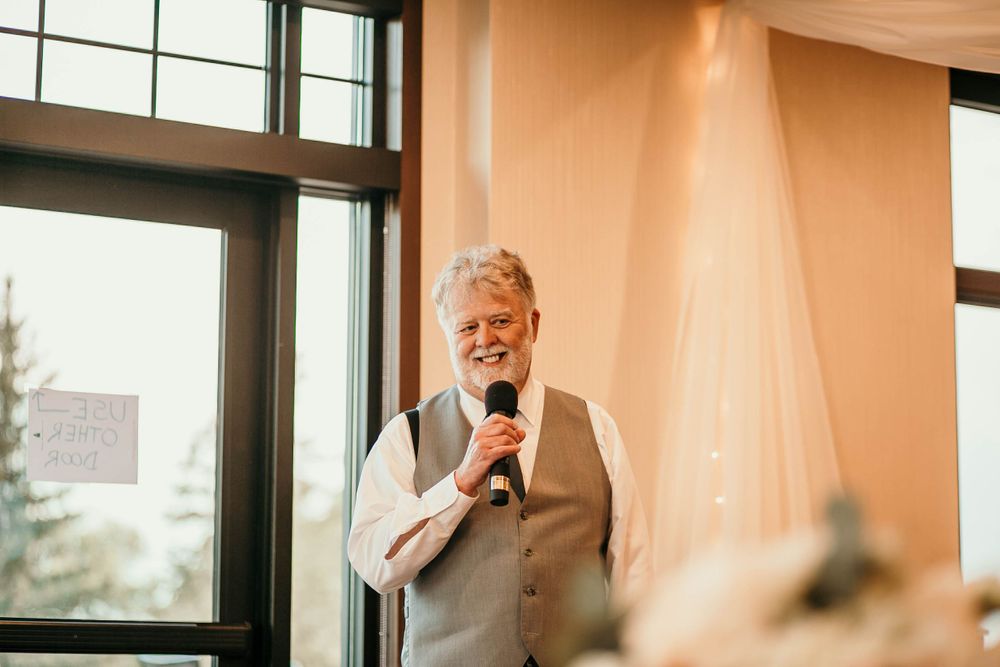 Older gentleman in gray vest delivers heartfelt speech with microphone at elegant wedding reception.