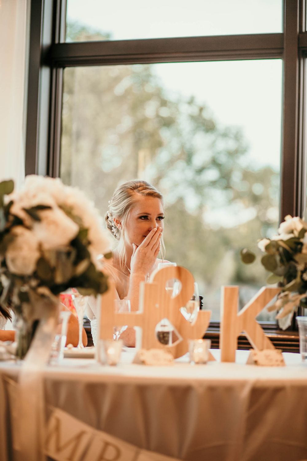 Emotional bride wipes tear at head table beside monogram letters and white floral centerpieces during speeches.