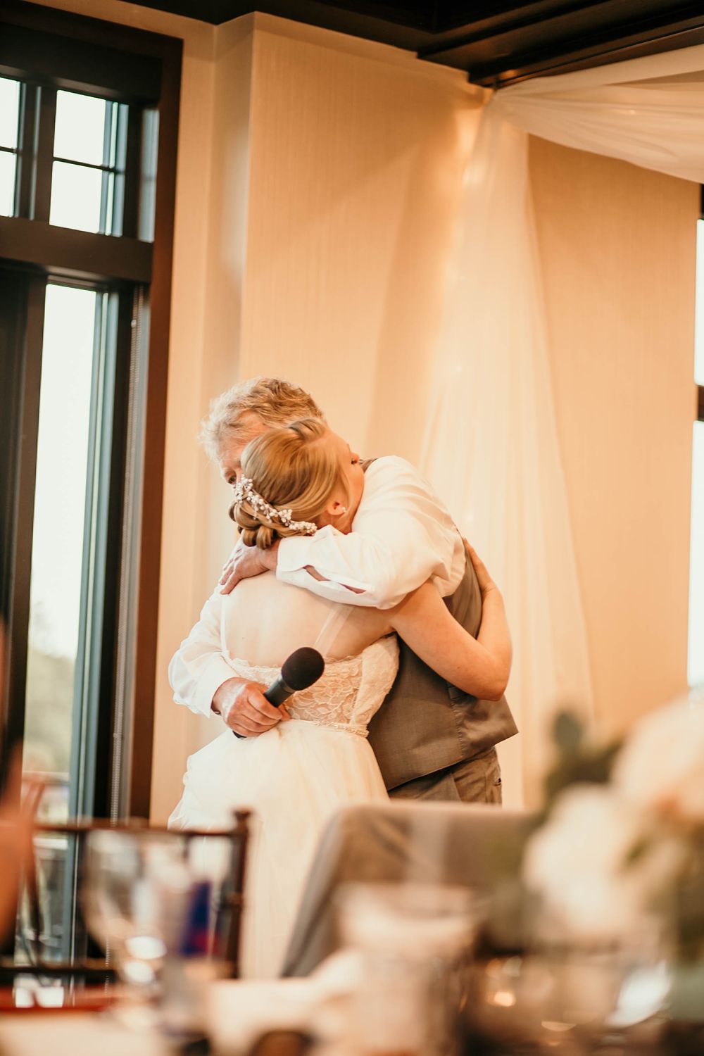 Bride in white gown shares emotional hug with older man holding microphone at wedding reception.