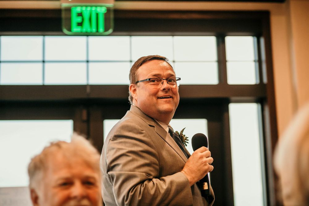 Man in a tan suit holding a microphone and smiling while giving a toast at a wedding reception.