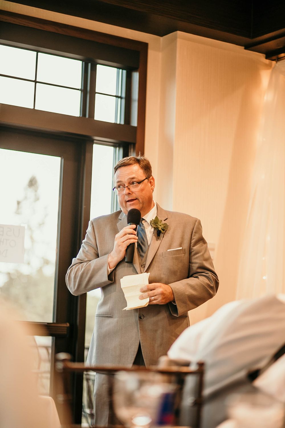 Man in gray suit holding a microphone and cup while giving a speech at a wedding reception.