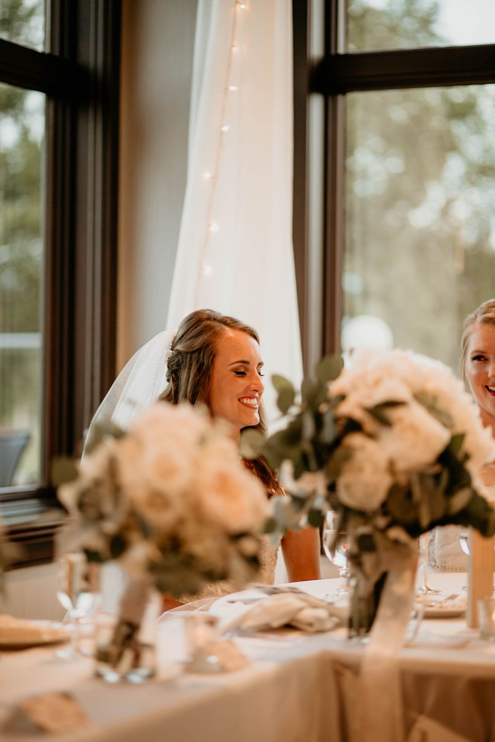 Smiling bride wearing a veil seated at a wedding reception table with white floral centerpieces.