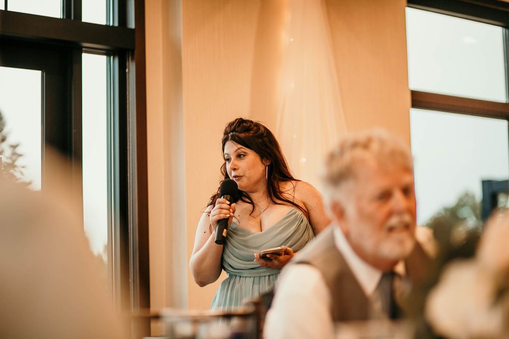Woman in blue bridesmaid dress speaking into a microphone during wedding reception toasts.