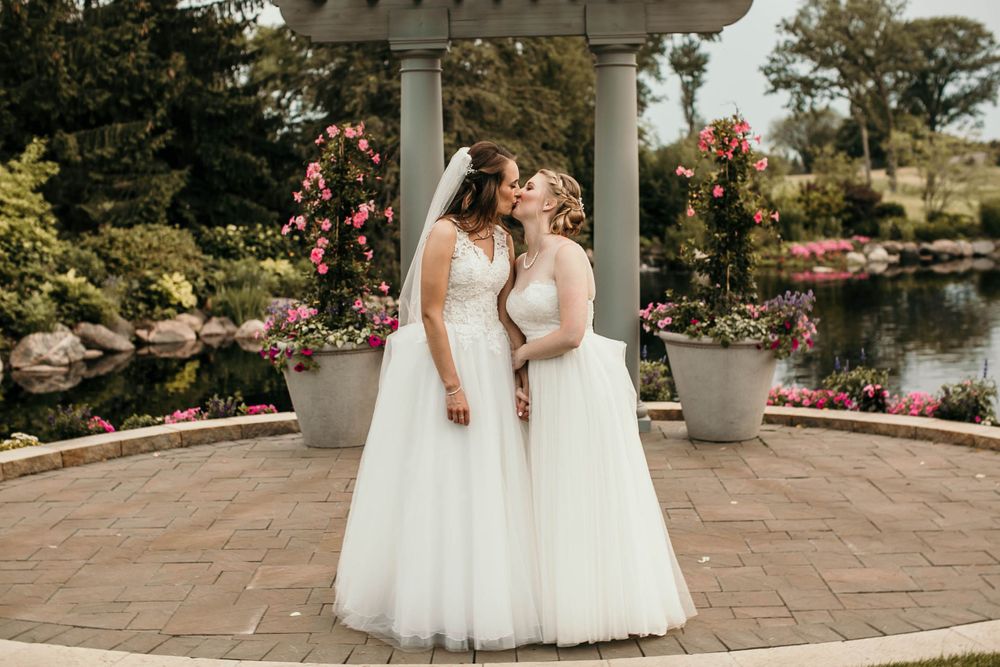 Two brides in white wedding gowns holding hands and sharing a tender moment in a garden gazebo.