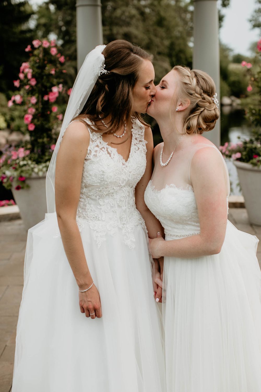Two brides sharing a romantic kiss outdoors, surrounded by blooming roses at their wedding.