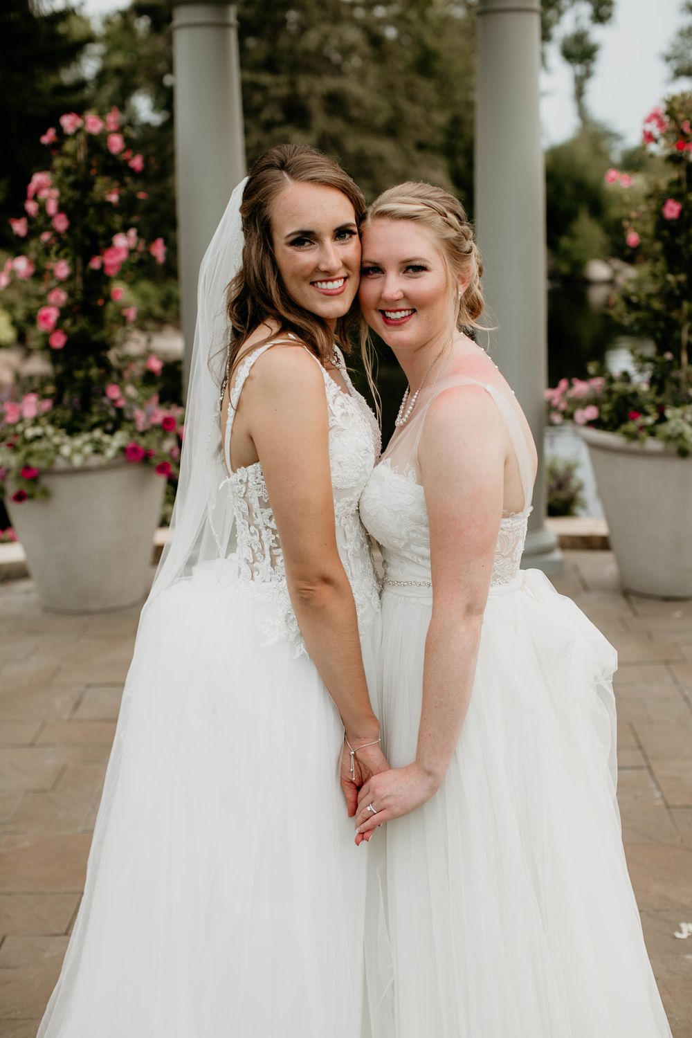 Two smiling brides in white wedding gowns holding hands and posing together in a rose garden.