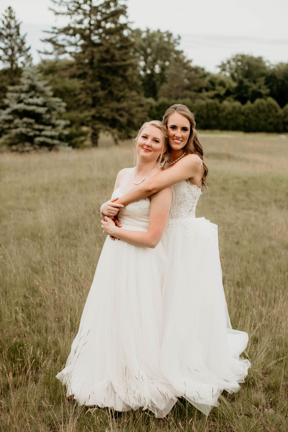 Two brides in elegant white wedding gowns share a loving embrace in a lush green meadow.