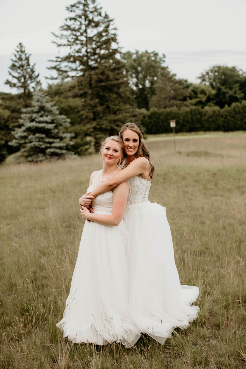 Two brides in white gowns embracing in an open field with evergreen trees in the background.