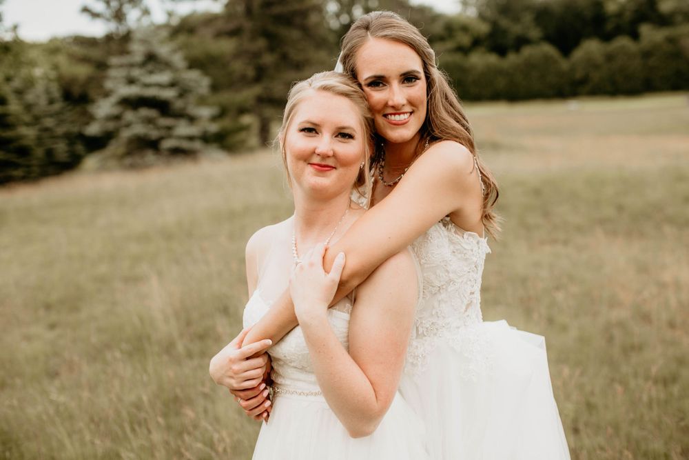 Two brides in white gowns embrace in a grassy field, smiling joyfully at their wedding.