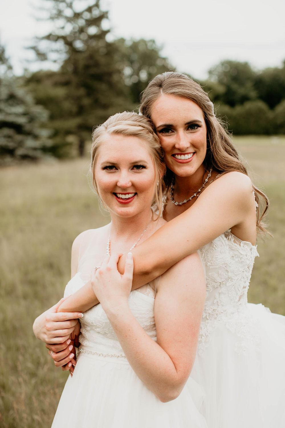 Two smiling brides in white lace wedding dresses hug warmly in a sunlit grassy field.