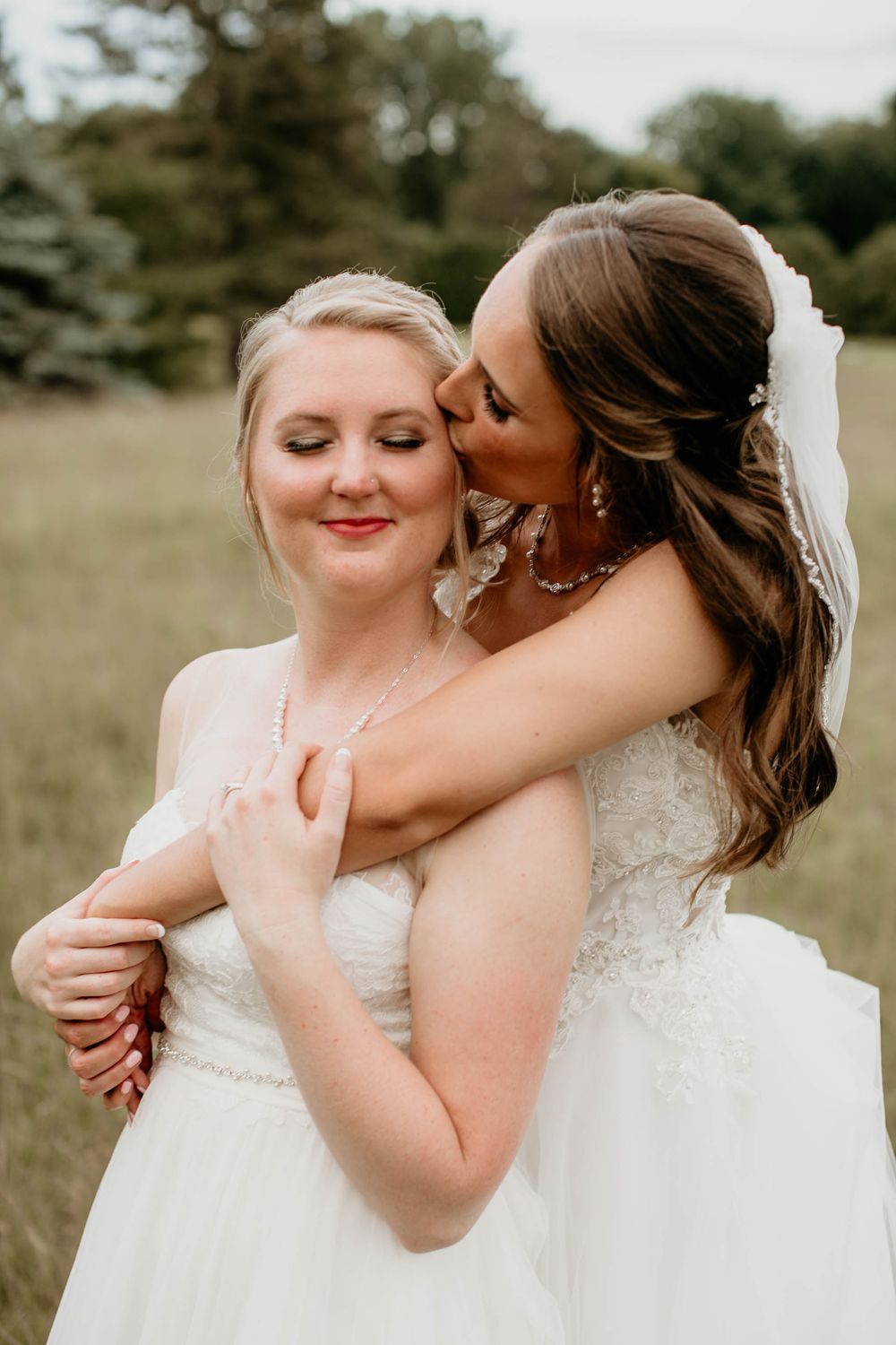 Two brides in white wedding gowns share an intimate moment, one whispering to the other outdoors.