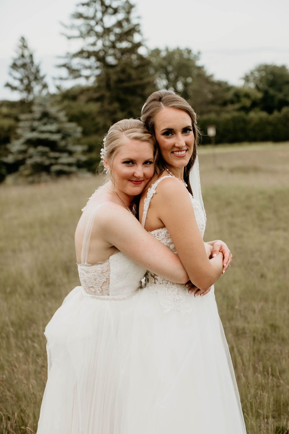 Two brides in white gowns embrace back-to-back, smiling in a scenic grassy field.