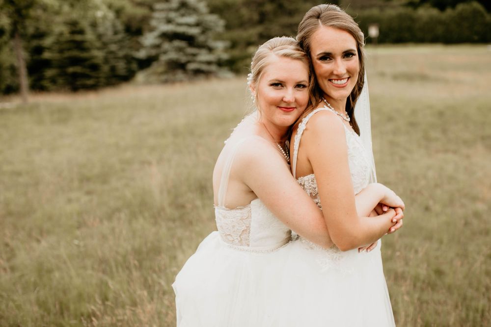 Two brides in white wedding dresses share a tender back-to-back embrace in a green meadow.