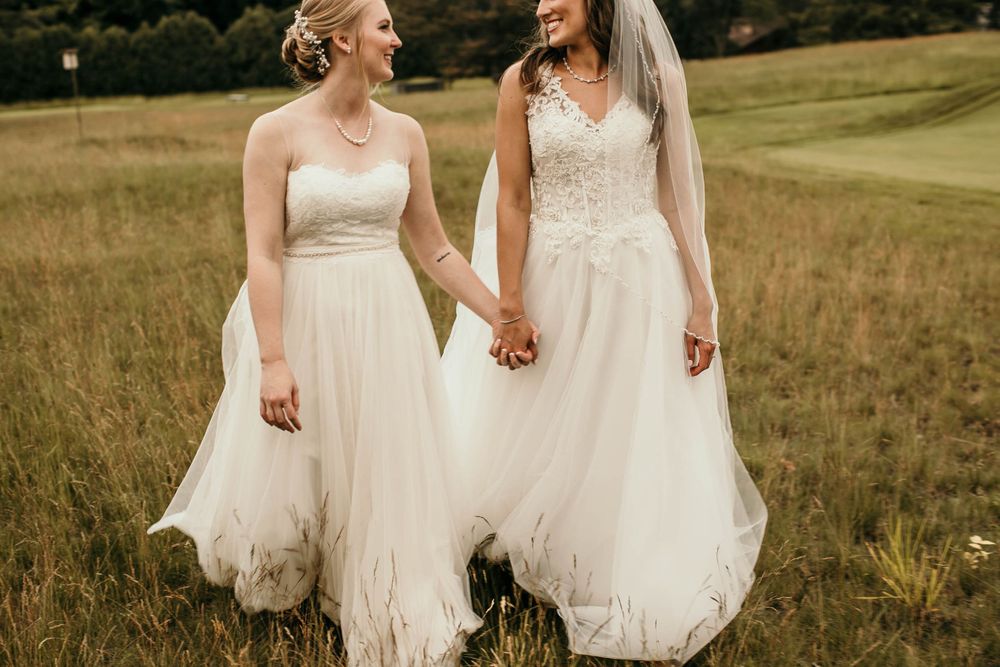 Two brides laugh and hold hands while walking through a lush green field in white gowns.