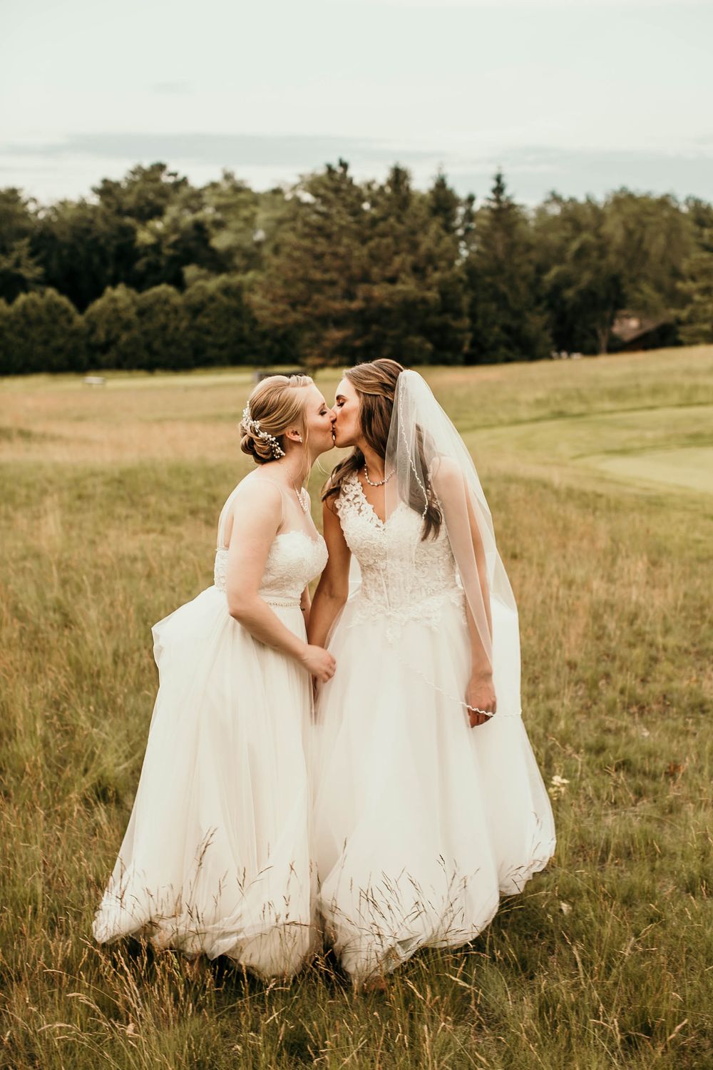 Two brides in white wedding gowns share a romantic kiss in a scenic open meadow.