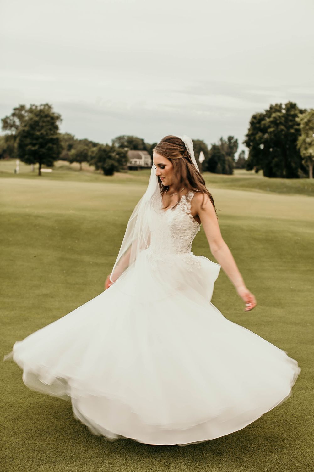 A bride in a white lace gown and veil twirls gracefully on a golf course green.