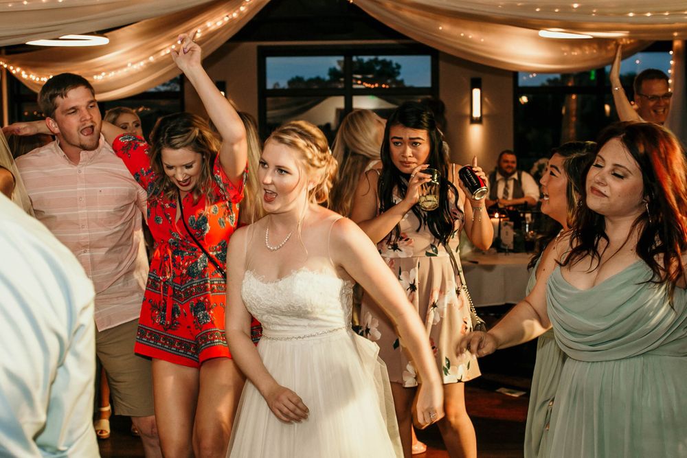 Bride in white gown dancing joyfully with wedding guests under string lights at reception.