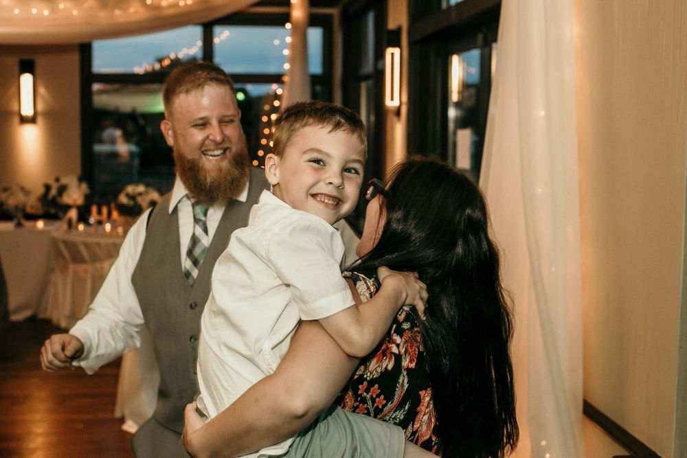 Smiling man in gray vest holds laughing young boy at wedding reception with string lights.