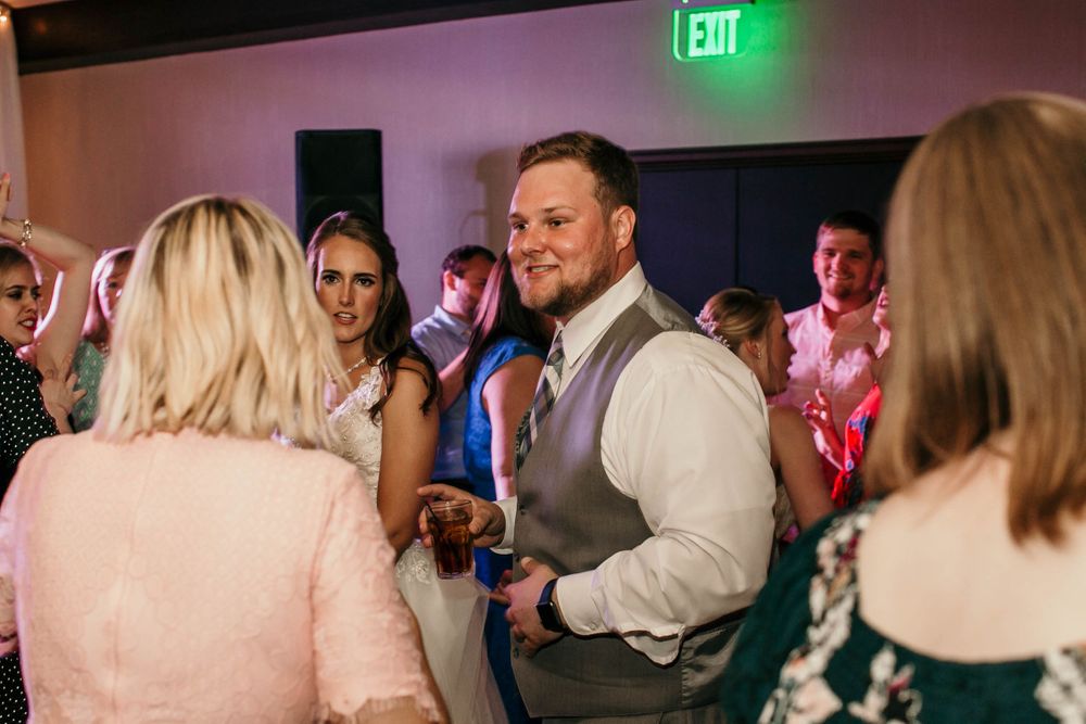 Groom in gray vest smiles while dancing with wedding guests on dimly lit reception floor.