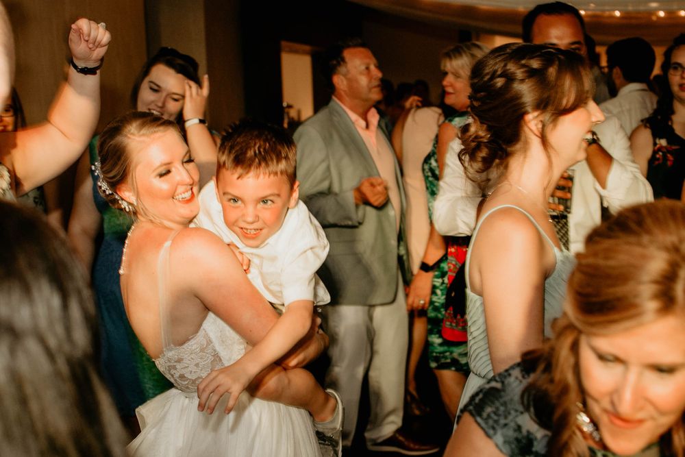 Bride in white gown hugs young boy while dancing with guests at wedding reception.