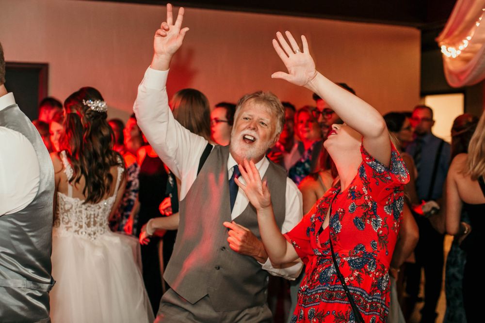 Excited wedding guests dance with hands raised under pink lights at lively reception party.