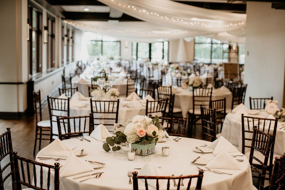 Bright wedding reception venue with round tables, dark Chiavari chairs, and draped string lights.