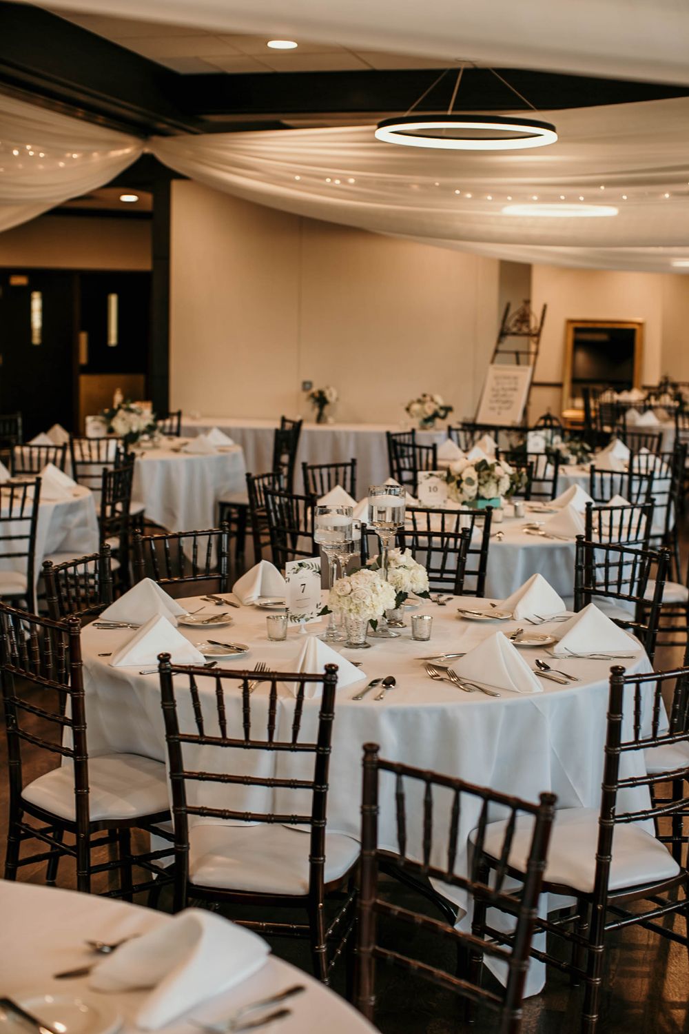 Elegant wedding reception hall with white tablecloths, dark Chiavari chairs, and circular chandelier.