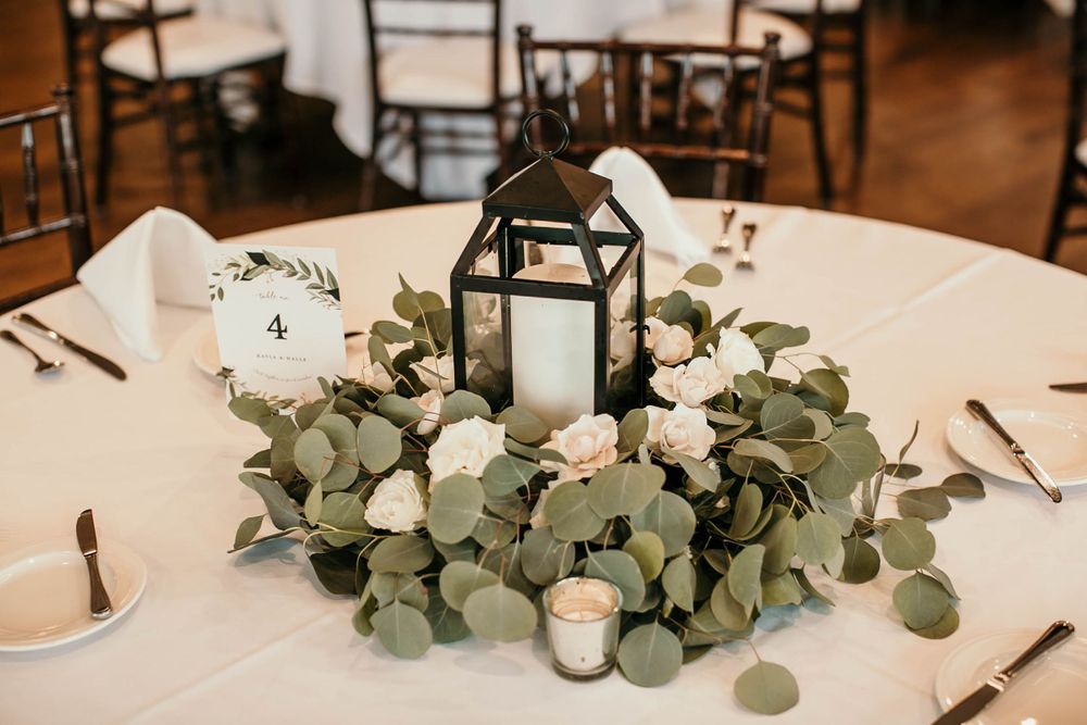 Elegant wedding centerpiece with black lantern, white roses, and eucalyptus wreath on white tablecloth.
